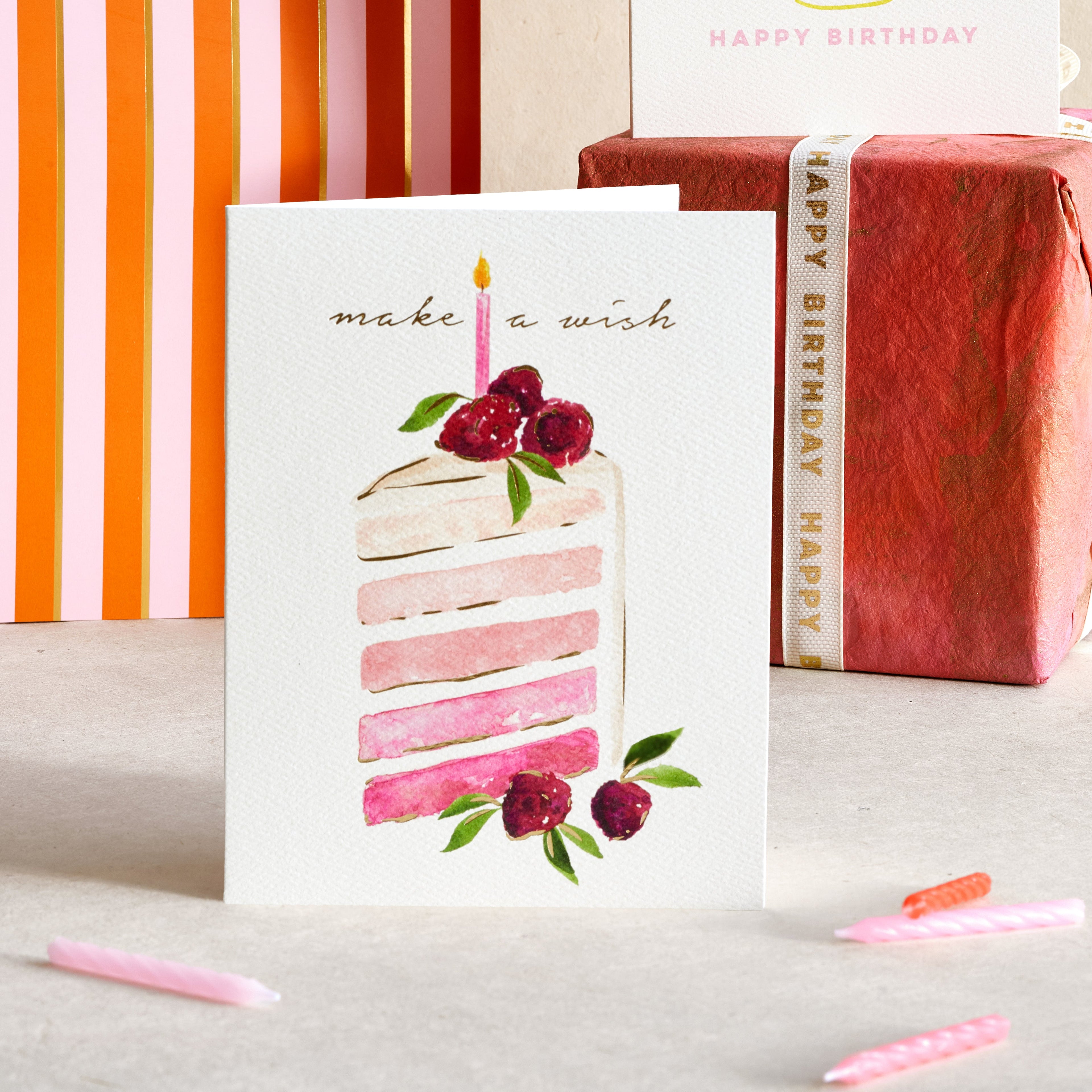 A birthday greeting card with a watercolor slice of pink cake and the words 'make a wish' stands in front of wrapped gift boxes and a striped bag. A second card in the background shows a pink cake and 'HAPPY BIRTHDAY' text.