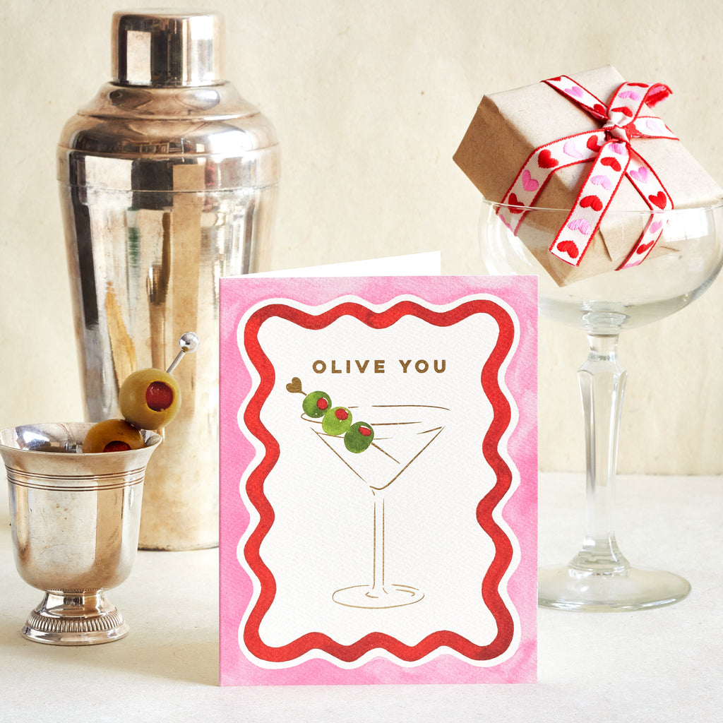 A greeting card with a pink border and an illustration of a martini glass with olives, reading 'OLIVE YOU', styled on a tabletop with a silver shaker, a glass, and a wrapped gift box.